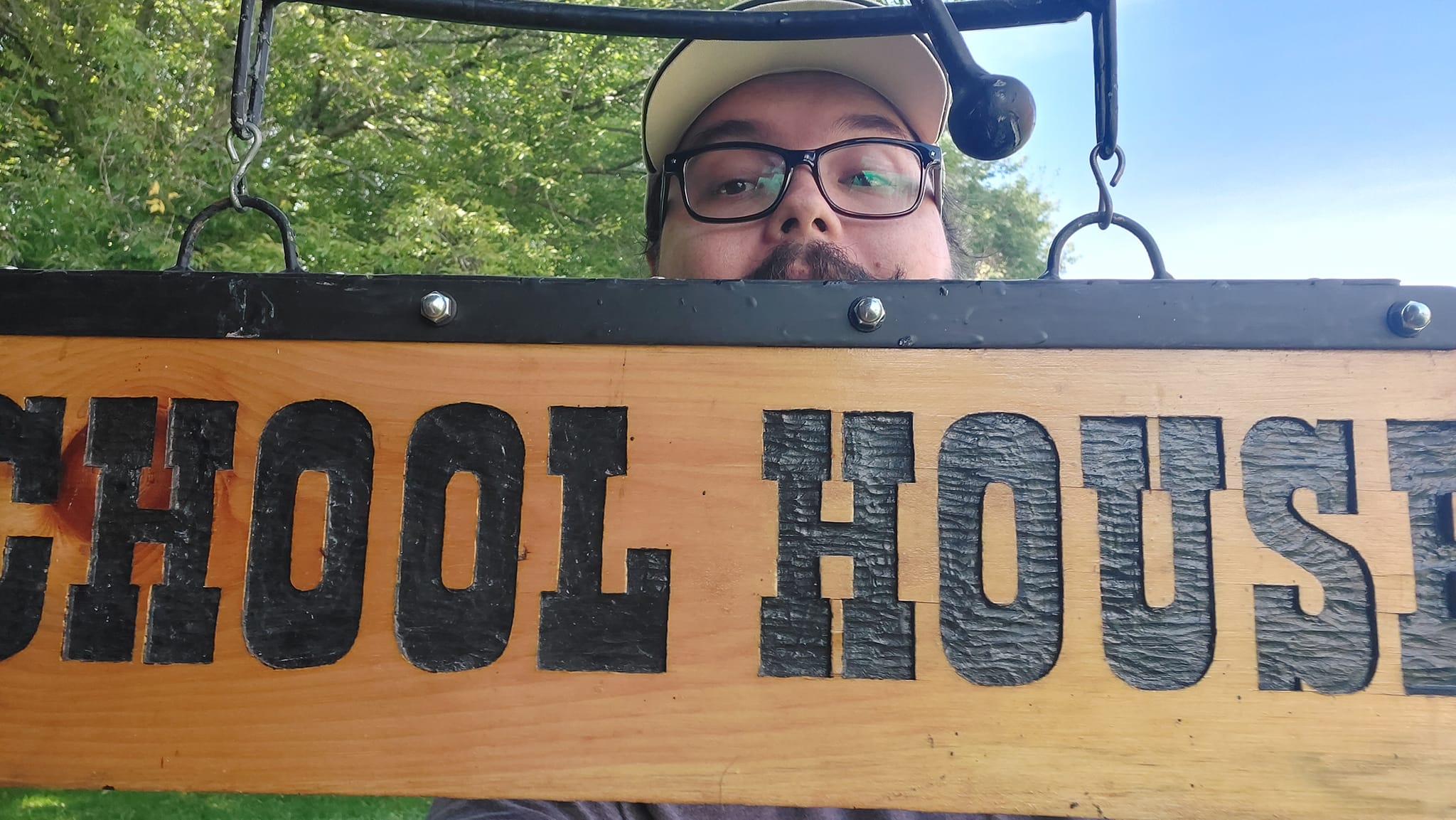 A man short hair and bushy facial hair pokes his head above a sign that reads "School House" done in an old style